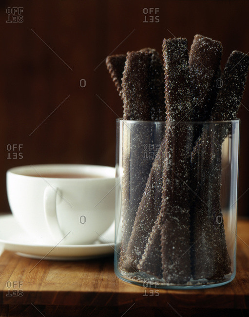 Dark brown baked sticks, sugared in clear glass vase with white coffee cup on wood surface with wood background