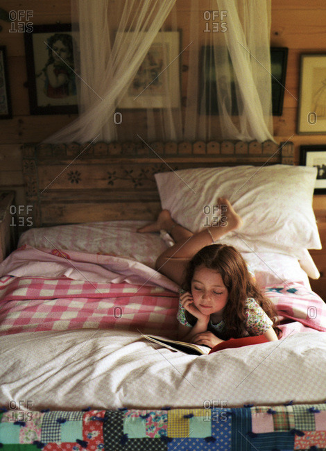 Young red headed girl reading on her stomach on her bed with vintage quilts and camp blankets and carved wooden headboard