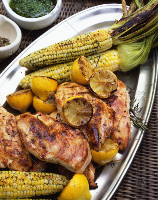 Grilled chicken breast with corn, halved lemons and rosemary on a metal serving tray.