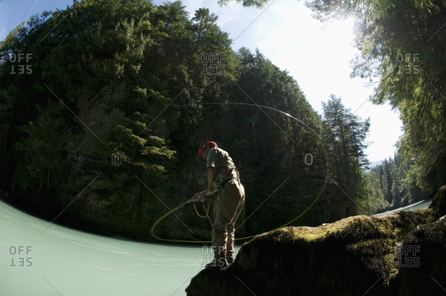 A man standing on a rock, casts into a river wearing waders while fly fishing in Squamish, British Columbia