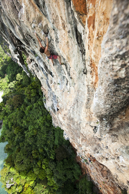 A athletic man rock climbing high above the trees and water on a multi pitch route in Thailand.
