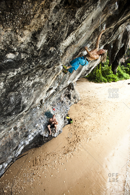 A athletic man rock climbing off the beach with his partner belaying from below in Thailand.