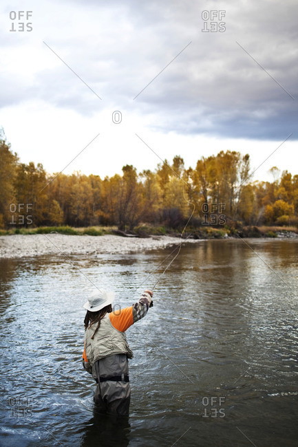A athletic man fly fishing stands in a river with the fall colors and snowy mountains behind him.