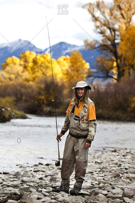 A athletic man fly fishing stands on the banks of a river with the fall colors and snowy mountains behind him in Montana.