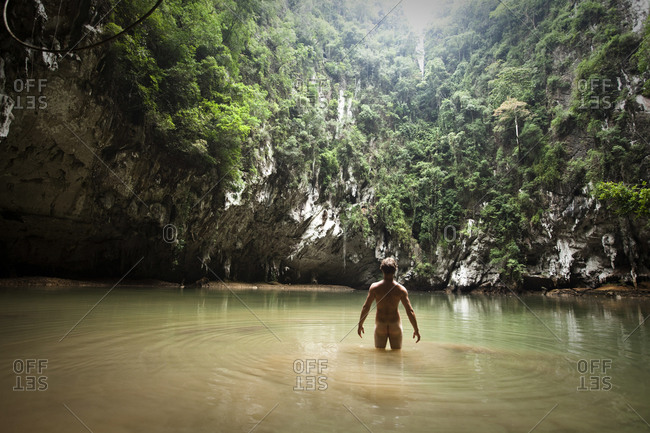 A athletic young man adventuring deep into a remote jungle pool stands naked in Thailand.