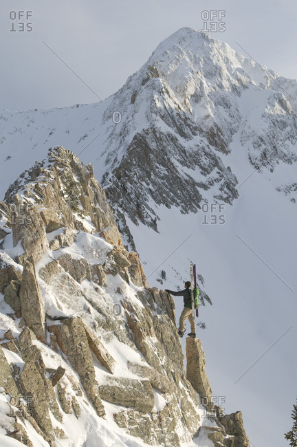 A man on a rocky ridge with skis below snowy mountain, Red Pine Canyon, Wasatch National Forest, Salt Lake City, Utah.