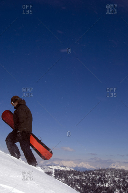 A young man walks up a snowy mountain with his snowboard in the backcountry
