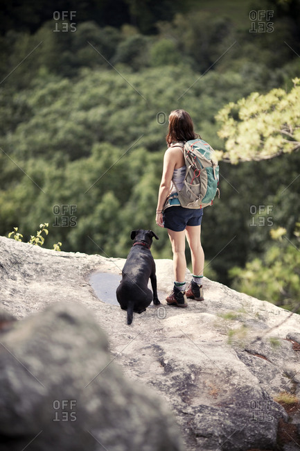 A female hiker and her dog look out over dense forest from a clifftop viewpoint.