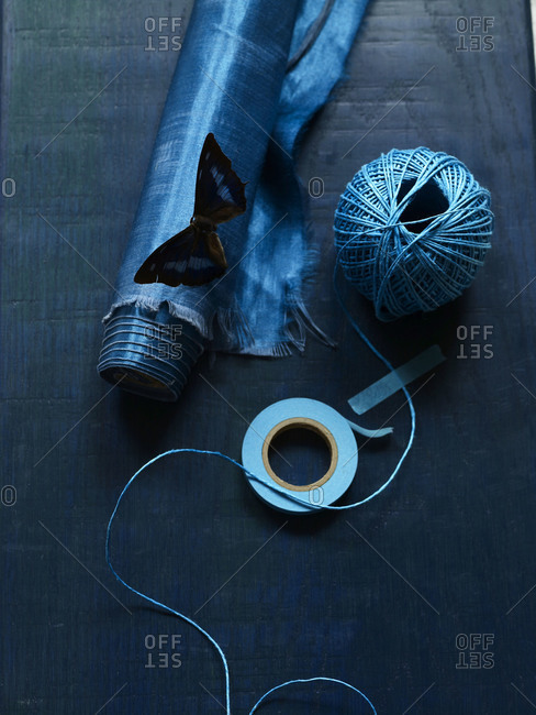 Still life of indigo blue roll of fabric, butterfly, decor tape and spool of string.