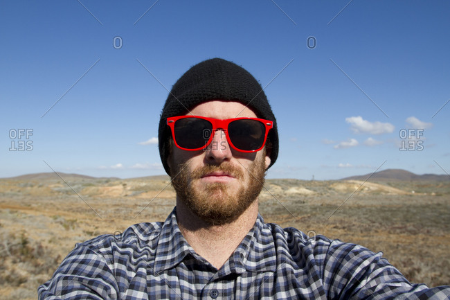 A male surfer with a beard wearing red sunglasses and a back beanie takes a self-portrait during a surf trip in Central Baja, Mexico.