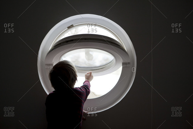 A young boy reaching to open a round window in silhouette