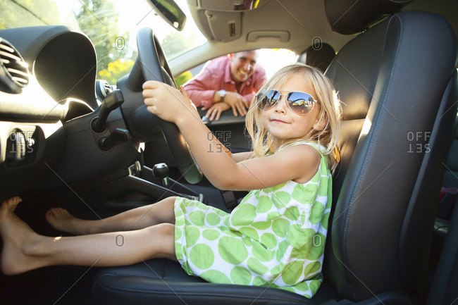Portrait of little girl sitting in driver\'s seat of car, pretending to be old enough to drive as her smiling father watches on on a sunny summer evening in Portland, Oregon, USA