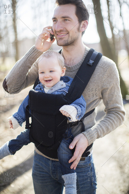 Happy young man using mobile phone while carrying baby boy in carrier at field