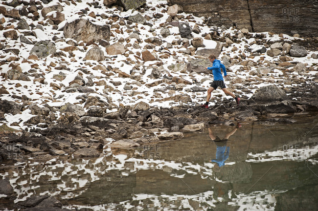 Athletic young man runs on a snowy mountain trail beside a lake with his reflection