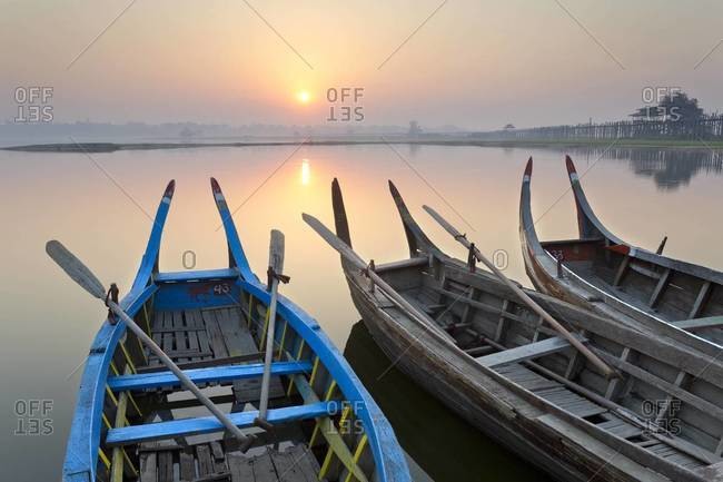 Traditional rowing boats moored on the edge of flat calm Taungthaman Lake at dawn with the  of the sky reflecting in the calm water, close to the famous U Bein teak bridge, near Mandalay, Myanmar (Burma), Asia