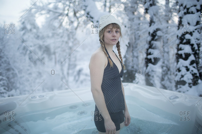 A young woman with a winter hat stands in a hot tub in the snowy forest