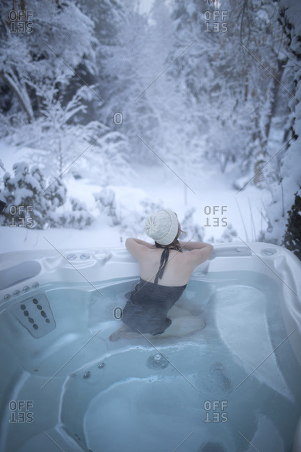 A young woman with a hat looks away into the snowy forest from a hot tub.