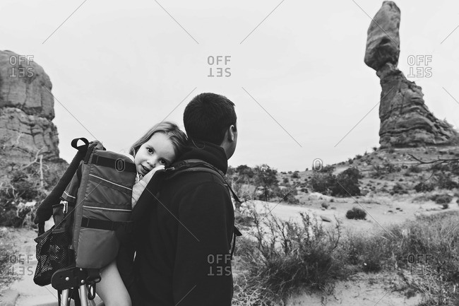 Black and white image of a father carrying his daughter on his back as he looks at Balanced Rock, Arches National Park, Utah