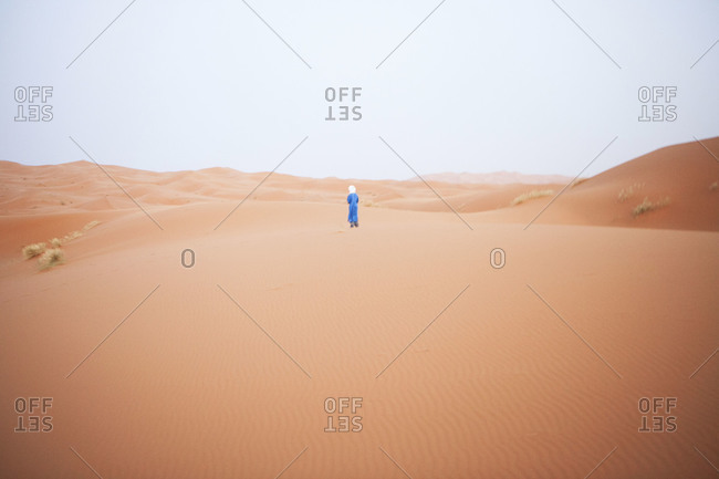 A solo Moroccan man dressed in traditional clothing walks alone in the Sahara desert