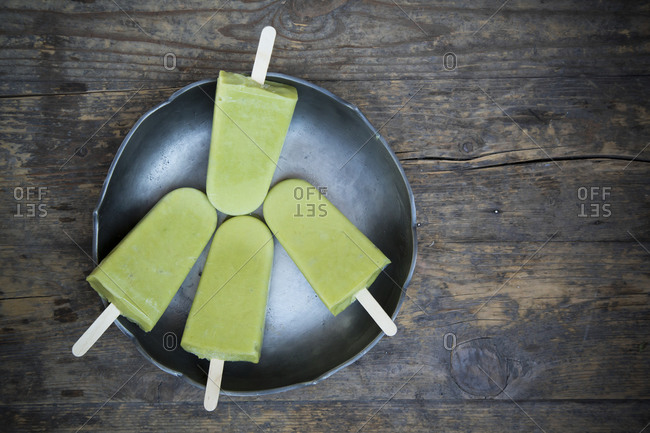 Bowl with four avocado ice lollies on dark wood, elevated view