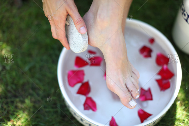 Woman taking an outdoor foot bath with rose leaves while exfoliating cracked skin on heel with pumice stone