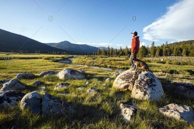 Man standing on a granite boulder with his dog with the Sierra Nevada mountains and the West Fork of the Carson River in the background in Hope Valley, California