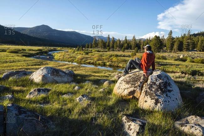 Man sitting on a granite boulder with the Sierra Nevada mountains and the West Fork of the Carson River in the background in Hope Valley, California
