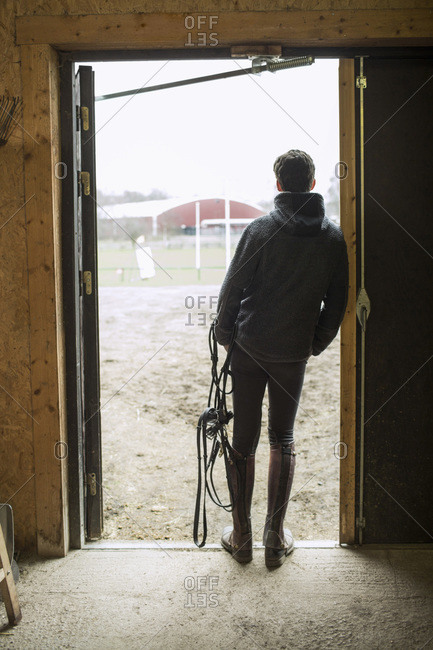 Rear view of young man with bridle leaning in doorway of horse stable