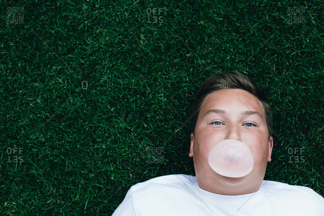 Teenage boy chewing bubble gum and blowing large bubble, laying on grass