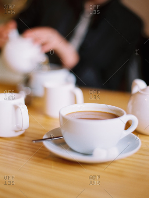 A woman seated pouring a cup of tea out of a white china tea pot.