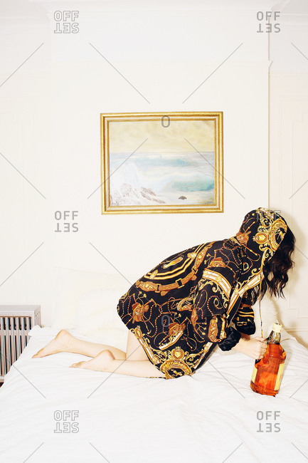 Woman in golden pattern hooded coat knees on bed with a bottle of alcohol