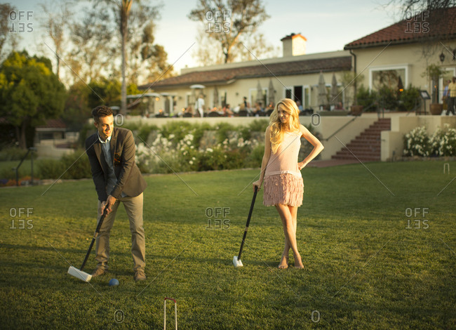 A smart young man hits a croquet ball with a mallet, a beautiful lady watches him from a distance with a mallet in her one hand and legs cross