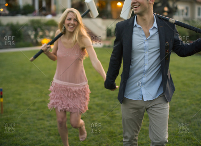 A young man with a croquet mallet on his left shoulder clutches the hand of his pretty young lady partner with a mallet on her right shoulder