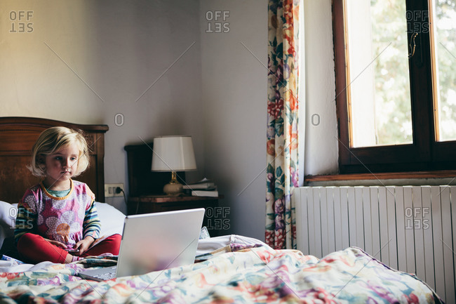 Cute three year old girl sitting on bed, watching movie on laptop computer