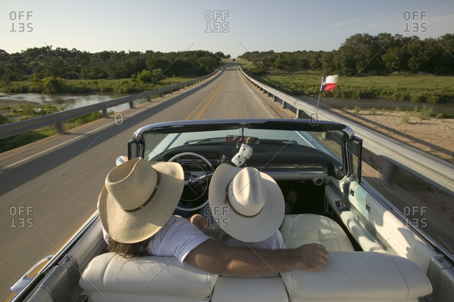 A couple takes a drive in their open top white car down Texas highways wearing cowboy hats