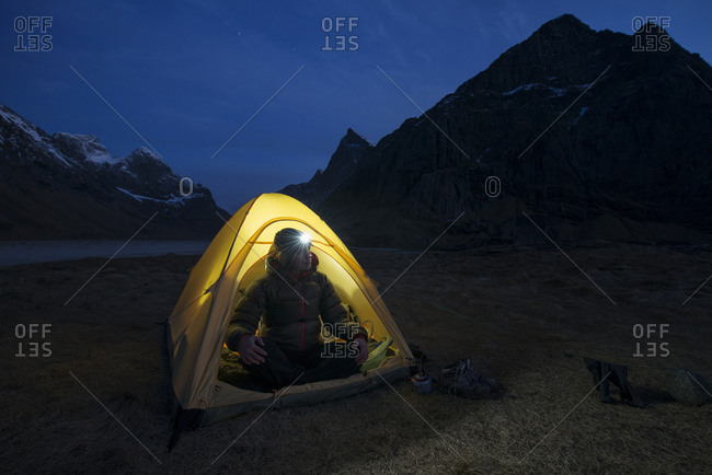 Female hiker sits in tent illuminated at night while wild camping at scenic Horseid beach, Moskenesøya, Lofoten Islands, Norway