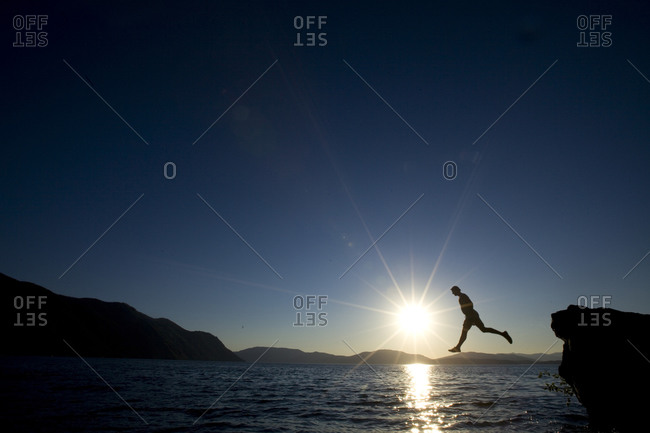 A man takes one big step off a rock into the water just before sunset