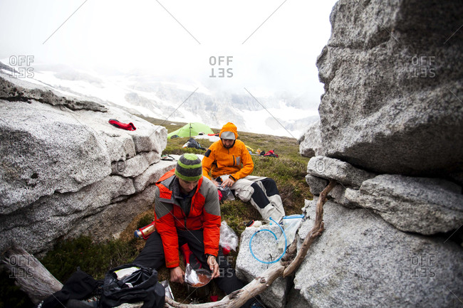 Two climbers prepare food while sitting between rocks hoping to avoid the wet conditions