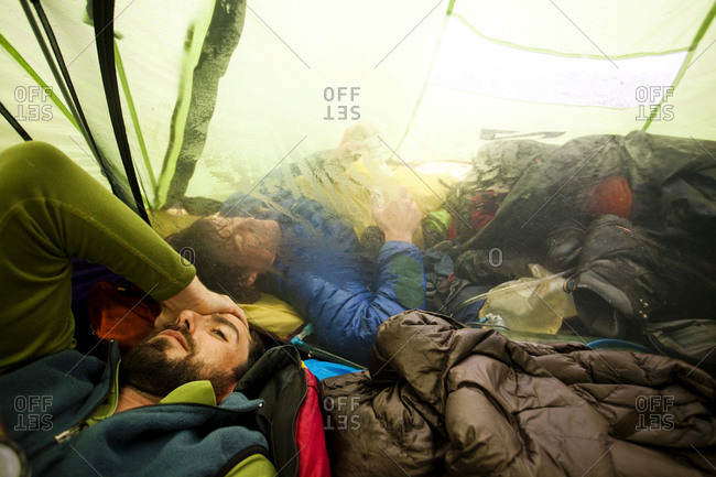 A climber rubs his head after a long day navigating through whiteout conditions while attempting to summit a mountain Another climber sleeps in the vestibule of the tent trying to avoid the rain