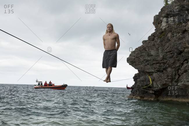 Male slackliner standing straight with arms folded behind the back on a 90 ft waterline while a patrol boat watches from a distance
