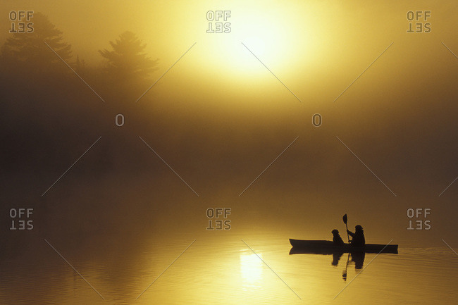 Silhouette of a woman sea kayaking with a dog at sunrise on Oxtongue Lake, Muskoka, Ontario, Canada