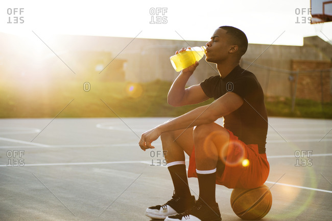 A man drinks a sports drink while sitting on a basketball