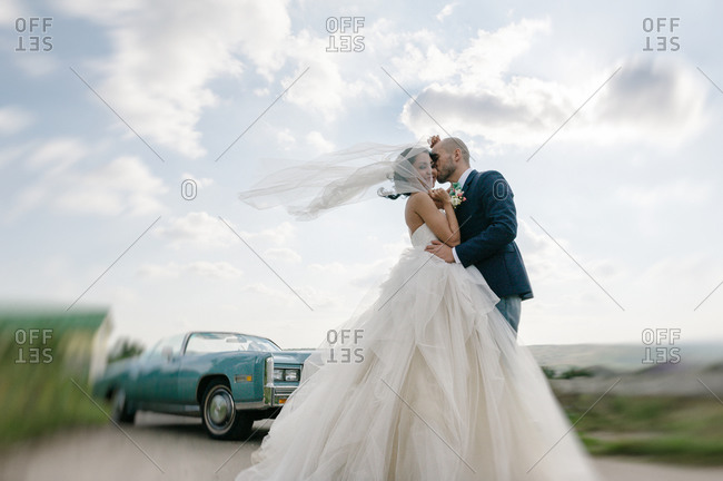Groom kissing bride in the middle of the road near a parked vintage car