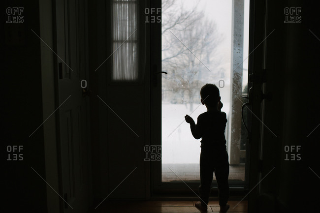 Boy standing silhouetted against glass door watching snow