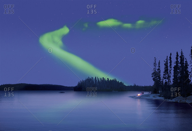 The Aurora borealis above a calm lake with two people by a fire on the lake shore