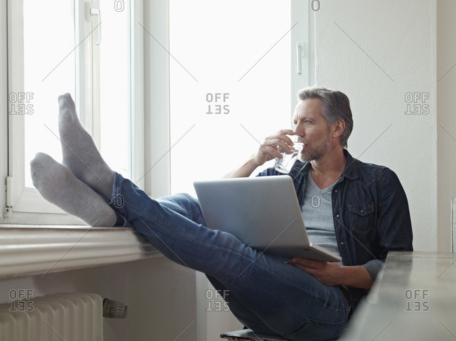 Mature man sitting at window using laptop with his feet up