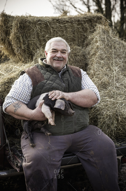 A large man, farmer in a waistcoat and working clothes, holding a piglet and smiling