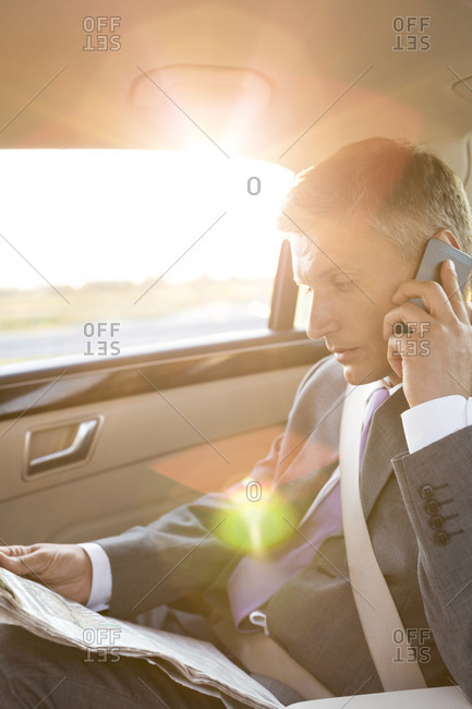 Businessman reading newspaper and using cell phone in car