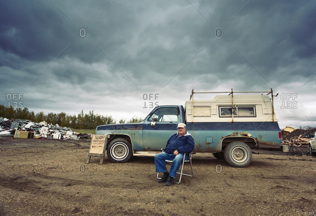 A mature man seated in a chair by his pick up truck surrounded by piles of waste, scrap metal and wooden objects