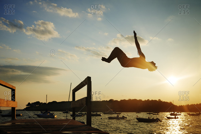 Teen boy doing back flip off pier at sunset